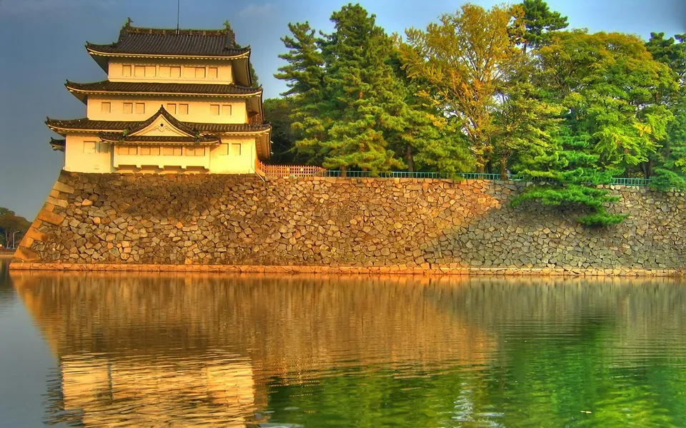 A multi-tiered Japanese castle with a stone base reflected in calm water, surrounded by trees.