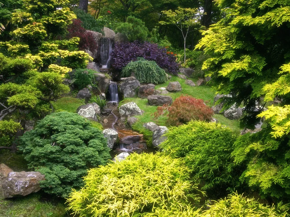 A serene Japanese garden with a multi-tiered waterfall flowing over rocks, surrounded by vibrant green and yellow trees and bushes.