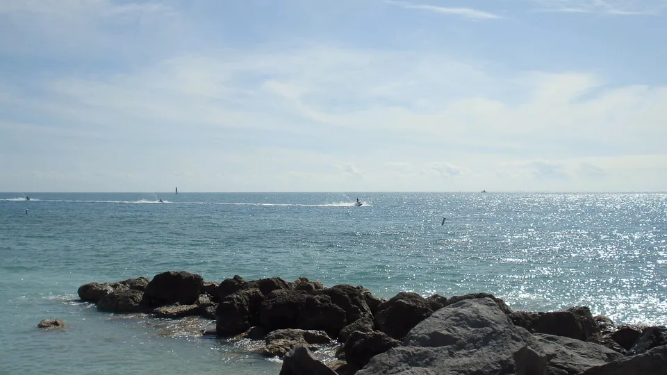 Jet skis ride on the ocean near a rocky shore under a partly cloudy sky.