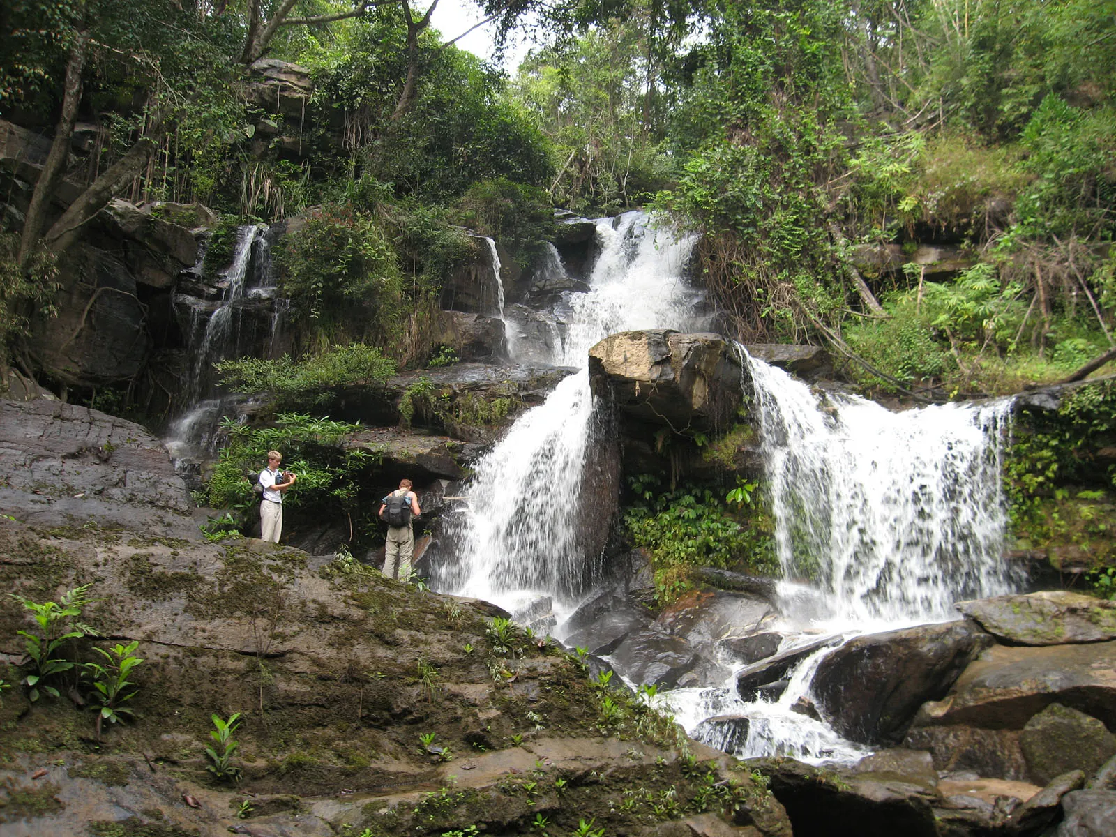 Two people stand on rocks near a multi-tiered waterfall cascading through a dense, green jungle.