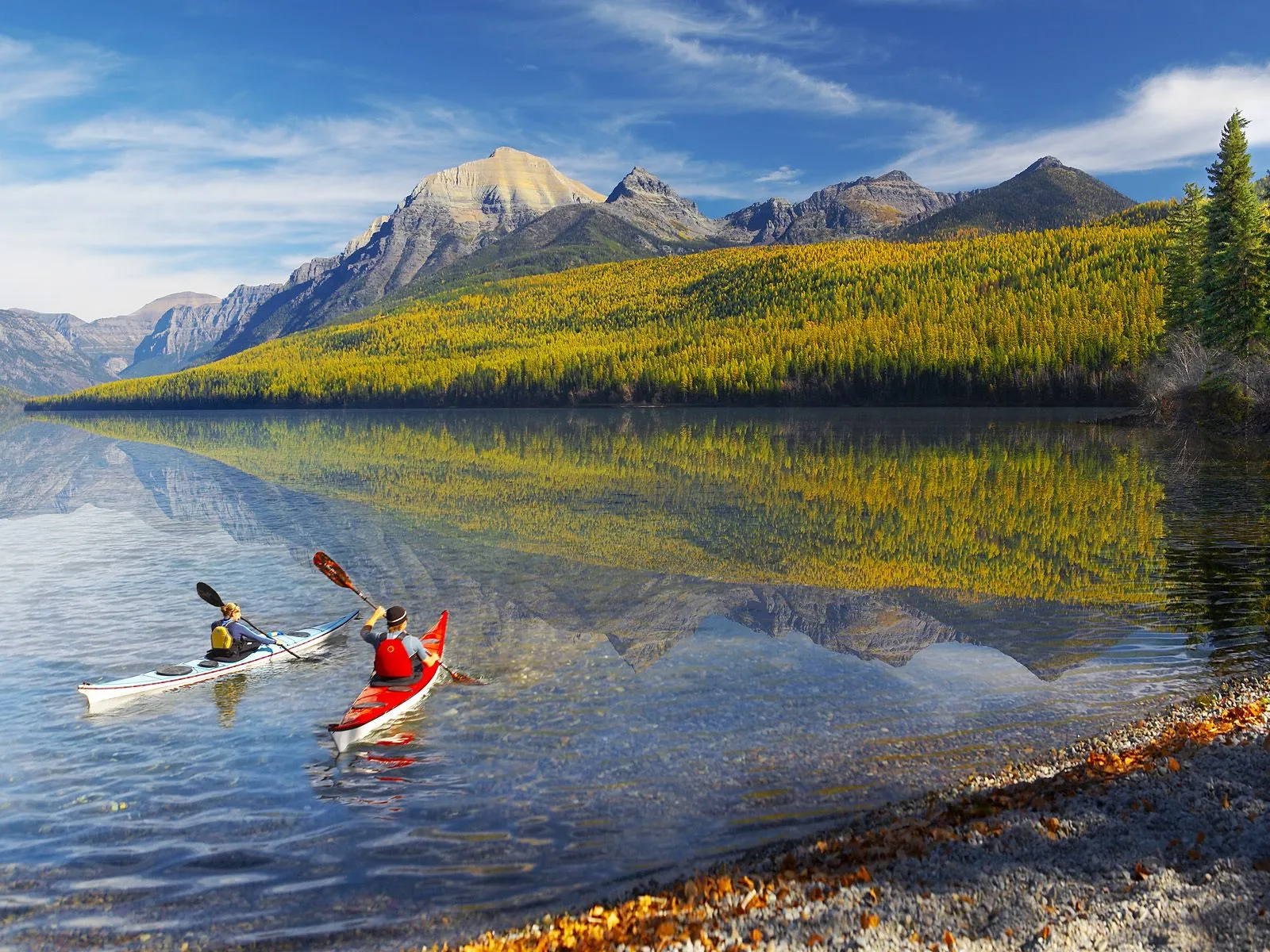 Two kayakers paddle on a clear, reflective lake with mountains and golden autumn trees in the background.