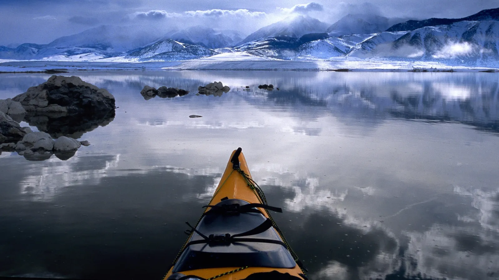 View from a yellow kayak looking out at a calm lake with snow-capped mountains in the background.