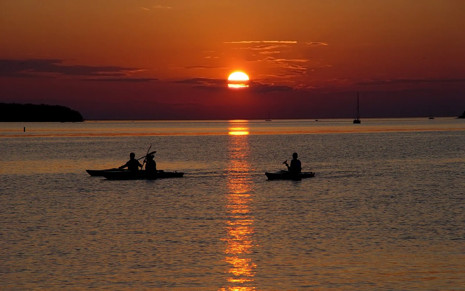 Two kayaks with people silhouetted against a vibrant orange sunset over the water.