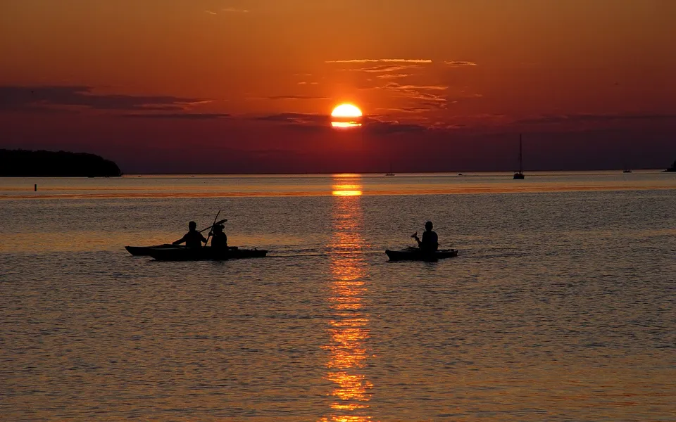 Two kayaks with people silhouetted against a vibrant orange sunset over the water.