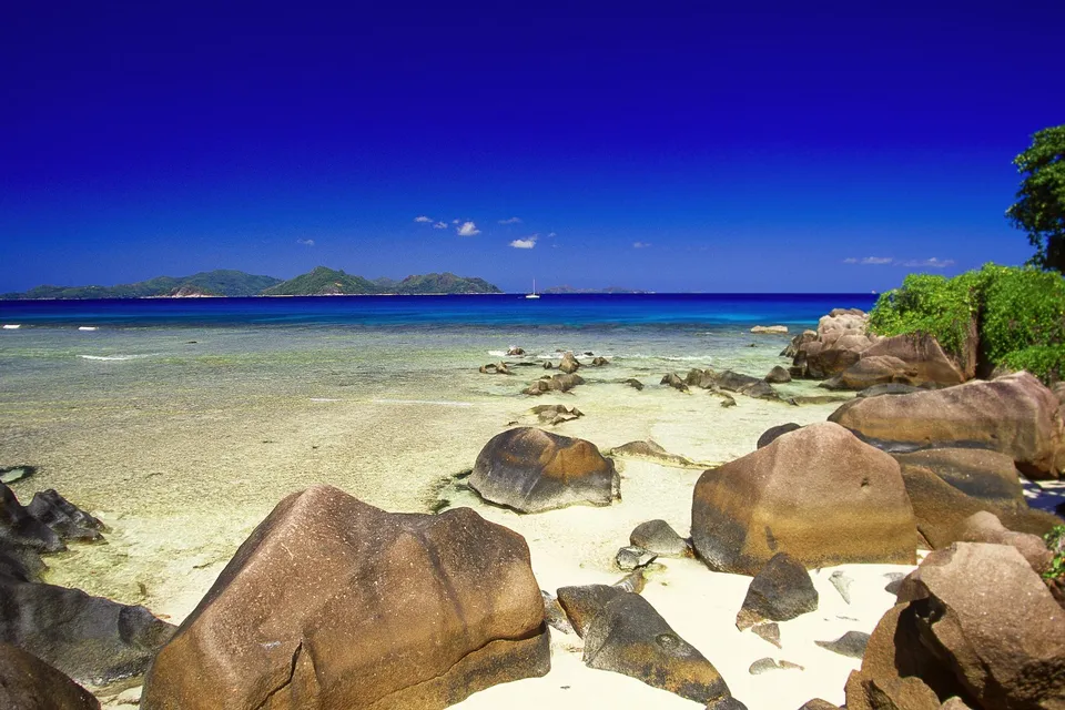 A tropical beach with large granite boulders, clear turquoise water, and lush green vegetation under a bright blue sky.