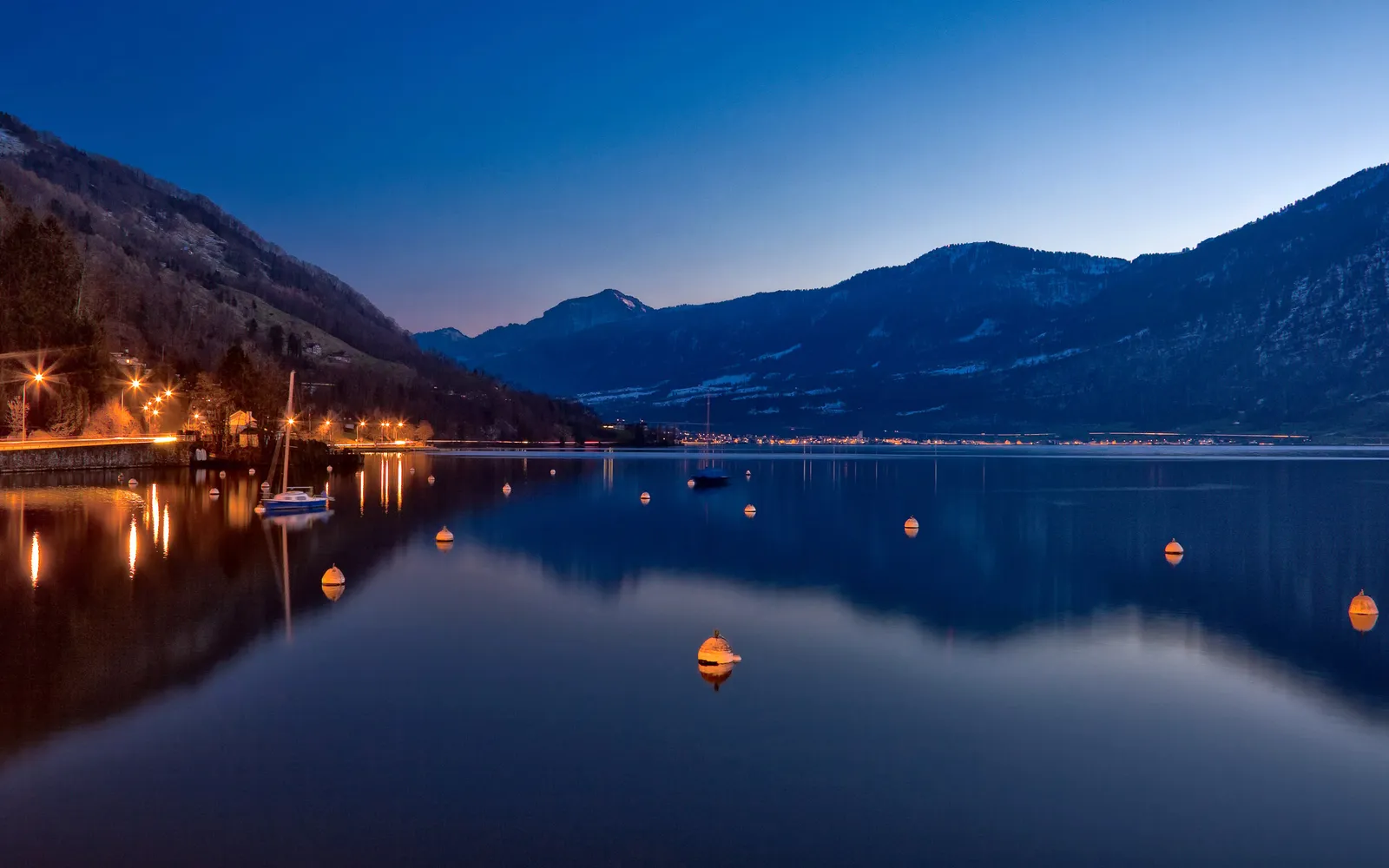 A calm lake reflects the twilight sky and distant mountains, with buoys and a sailboat visible.