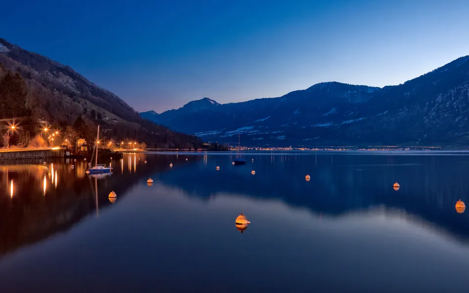 A calm lake reflects the twilight sky and distant mountains, with buoys and a sailboat visible.