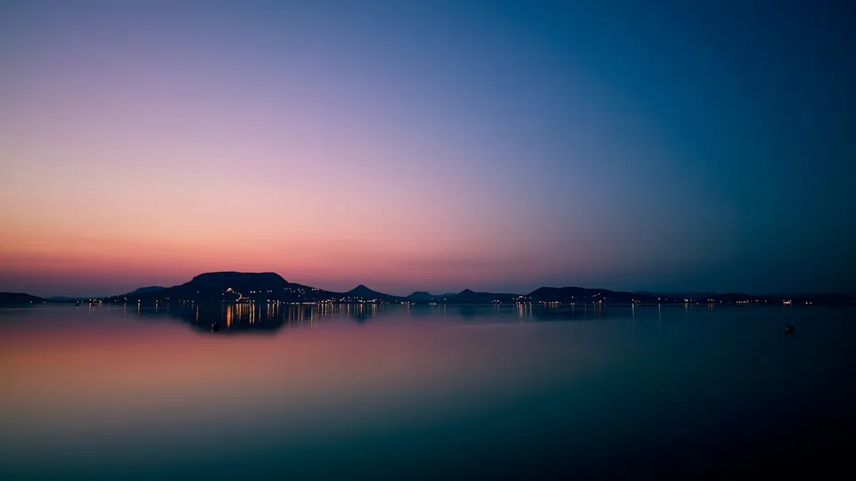A calm lake reflects the colorful sky and distant lights of a town on the shore at dusk.