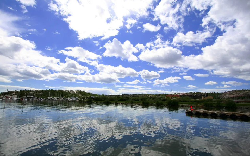 A wide shot of a calm lake with a marina and forested shoreline under a blue sky with white clouds.