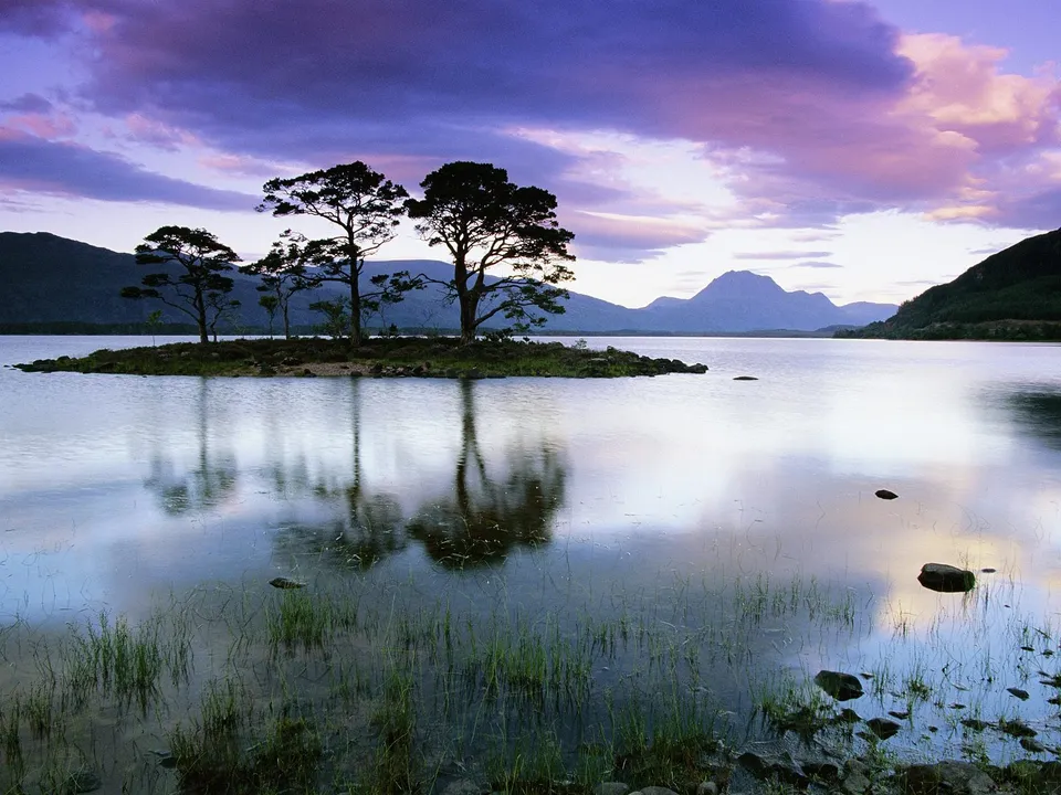 A serene lake reflects silhouetted trees on a small island under a dramatic purple and pink sky at dusk.