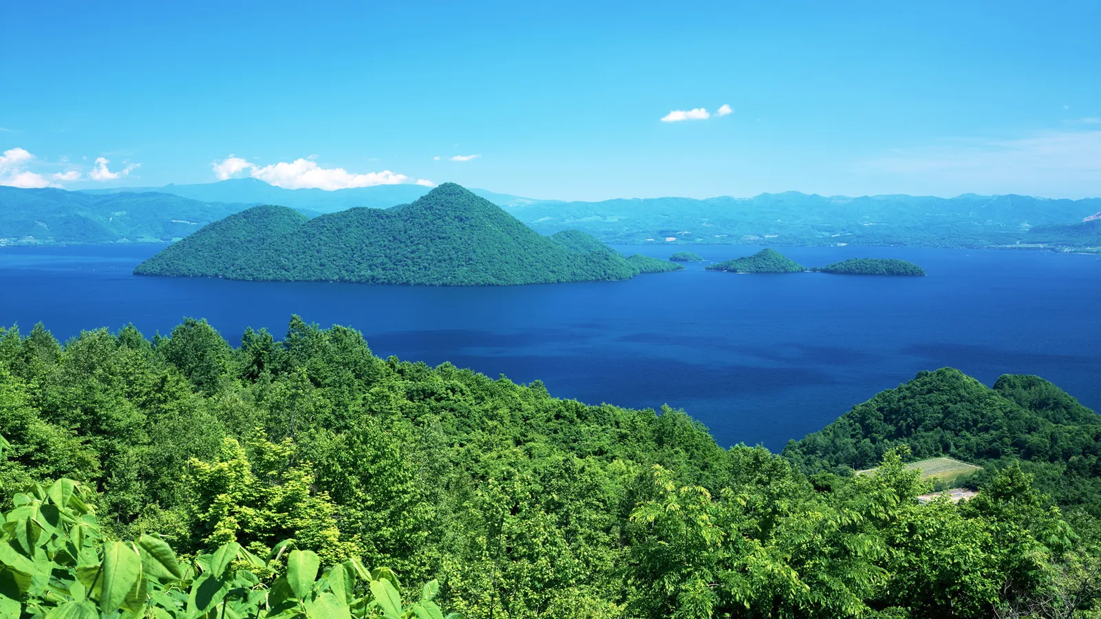 A wide view of a deep blue lake with tree-covered islands and distant mountains under a clear blue sky.
