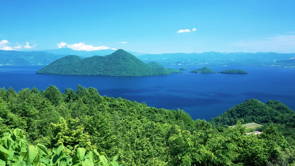 A wide view of a deep blue lake with tree-covered islands and distant mountains under a clear blue sky.