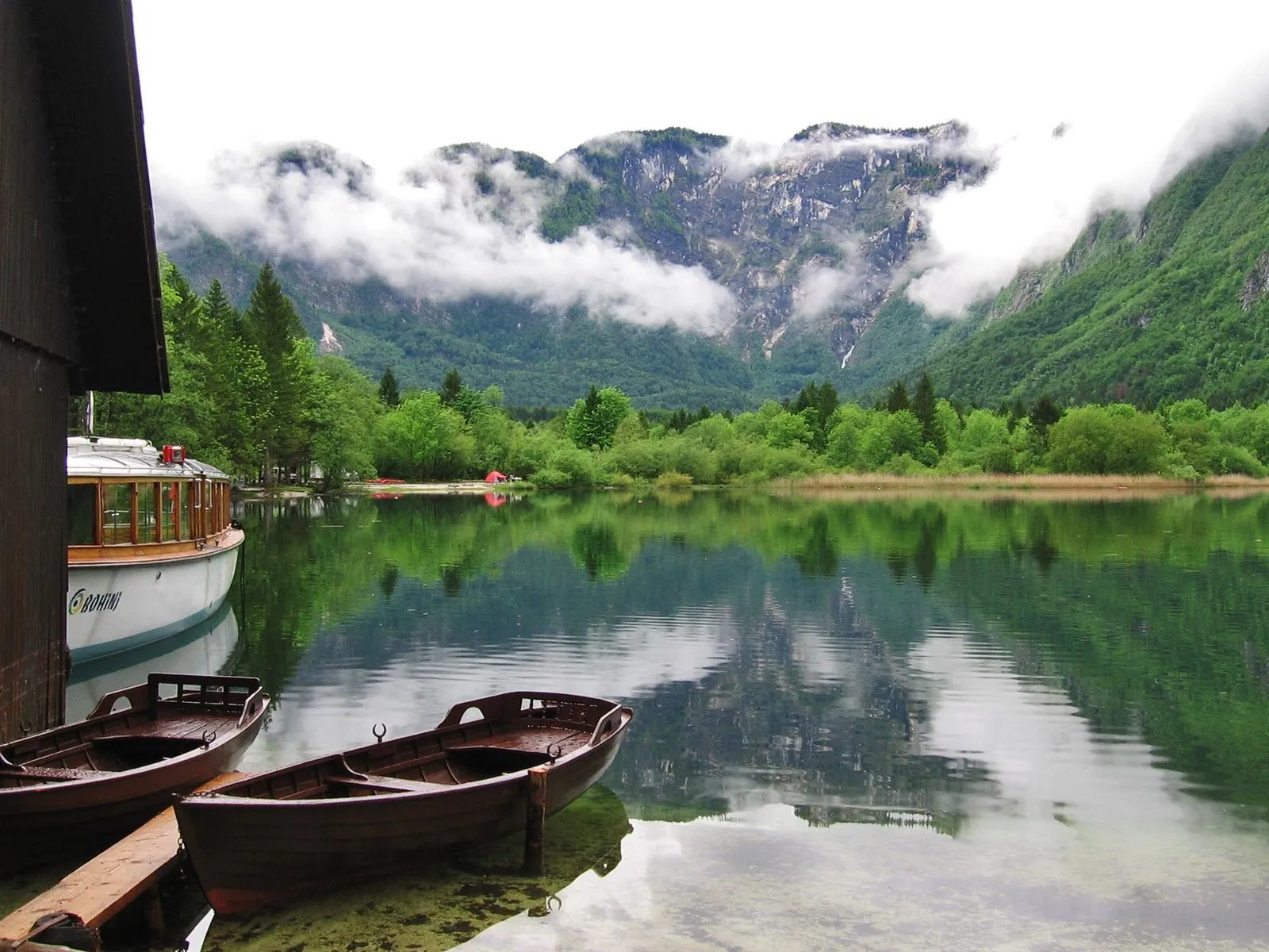 Two wooden rowboats are moored on the shore of a calm lake, with a large passenger boat docked nearby. Mountains are visible in the background.