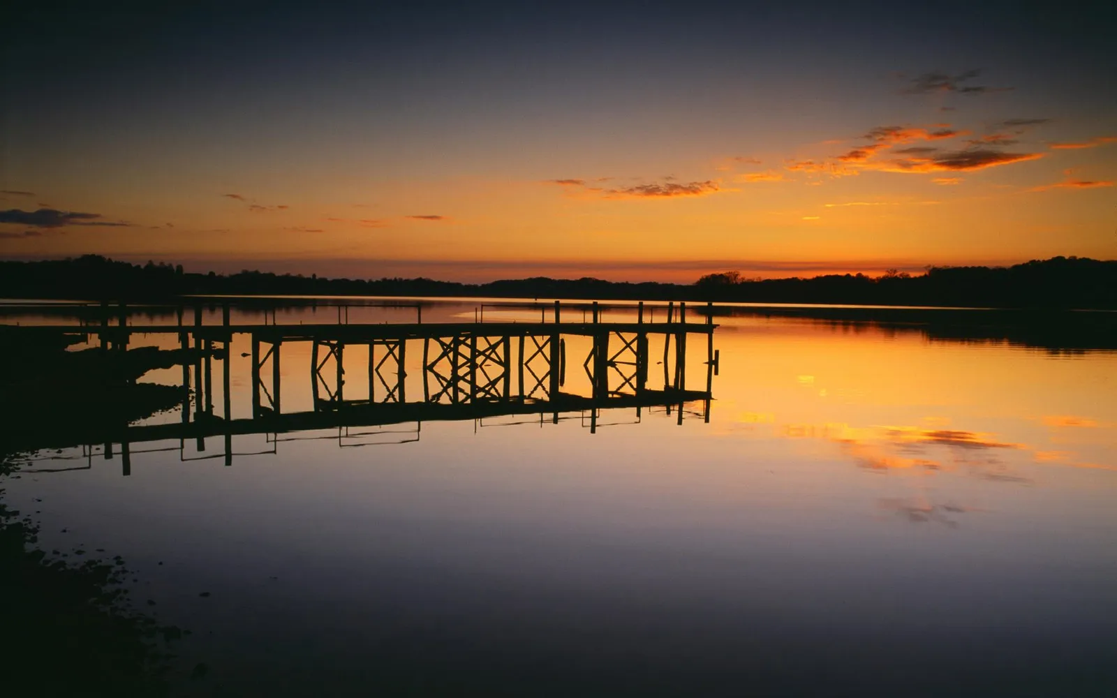 A silhouette of a wooden pier extends into a calm lake during a vibrant sunset.