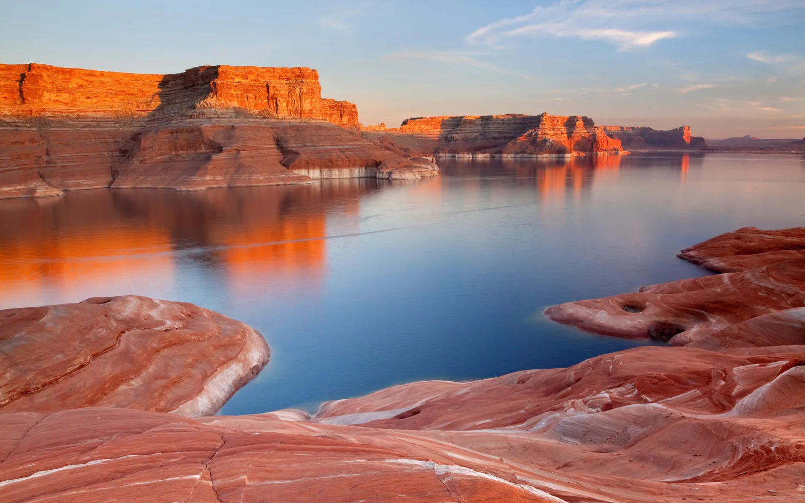 Red rock formations glow orange in the setting sun, reflected in the calm blue water of Lake Powell.
