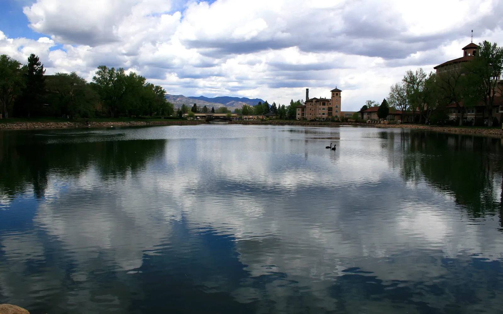 A calm lake reflects the sky with dramatic clouds and buildings on the shore.