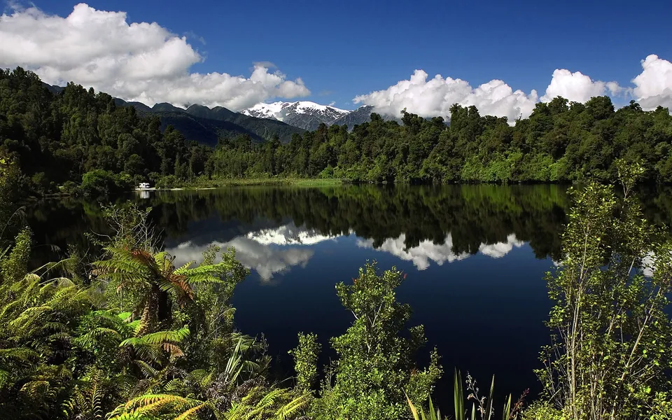 A tranquil lake reflects snow-capped mountains and a lush forest under a blue sky with white clouds.