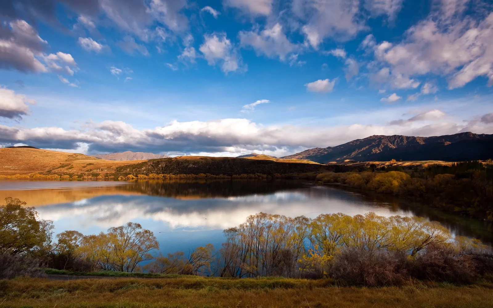 A calm lake perfectly reflects the sky and surrounding hills and trees.