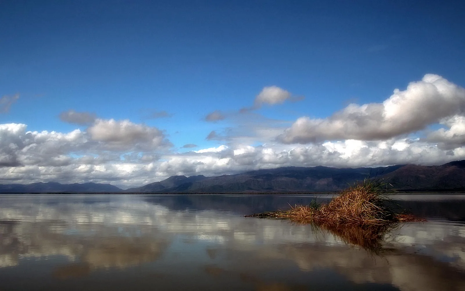 A calm lake reflecting a blue sky with scattered clouds and distant dark mountains.