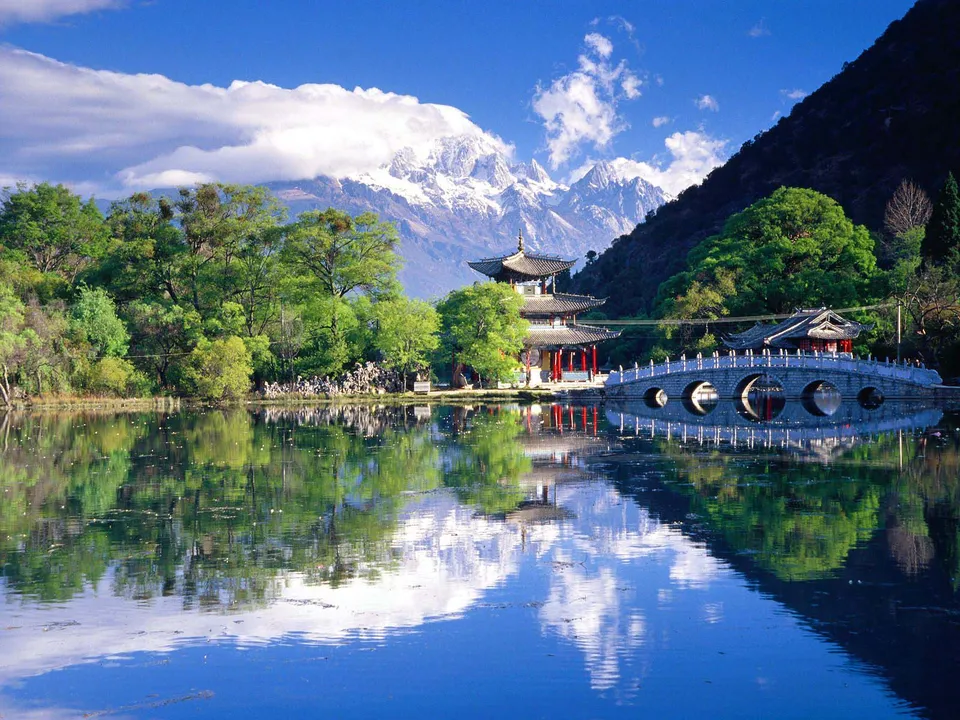 A tranquil lake reflects a traditional pagoda, arched bridge, lush trees, and snow-capped mountains under a blue sky.