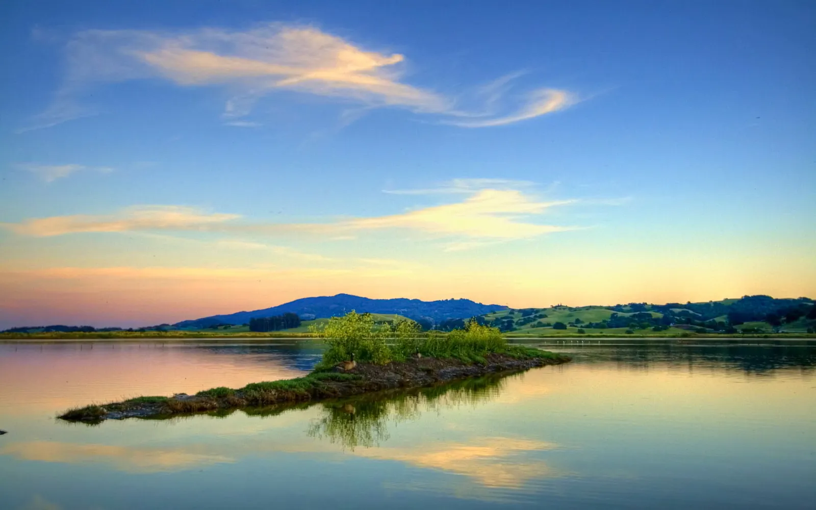 A small island with green vegetation sits in a calm lake, reflecting the soft colors of the sunset sky and distant hills.