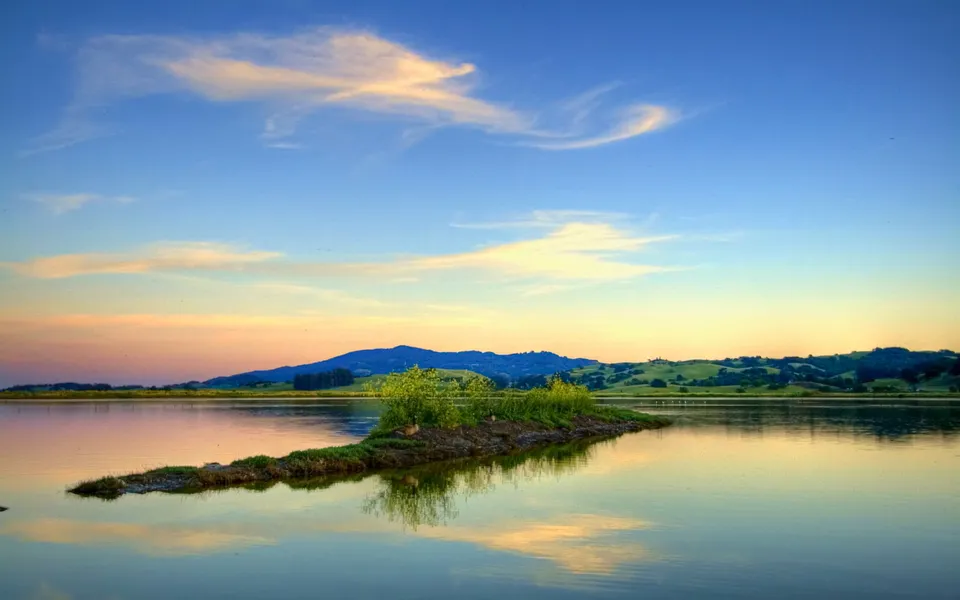 A small island with green vegetation sits in a calm lake, reflecting the soft colors of the sunset sky and distant hills.