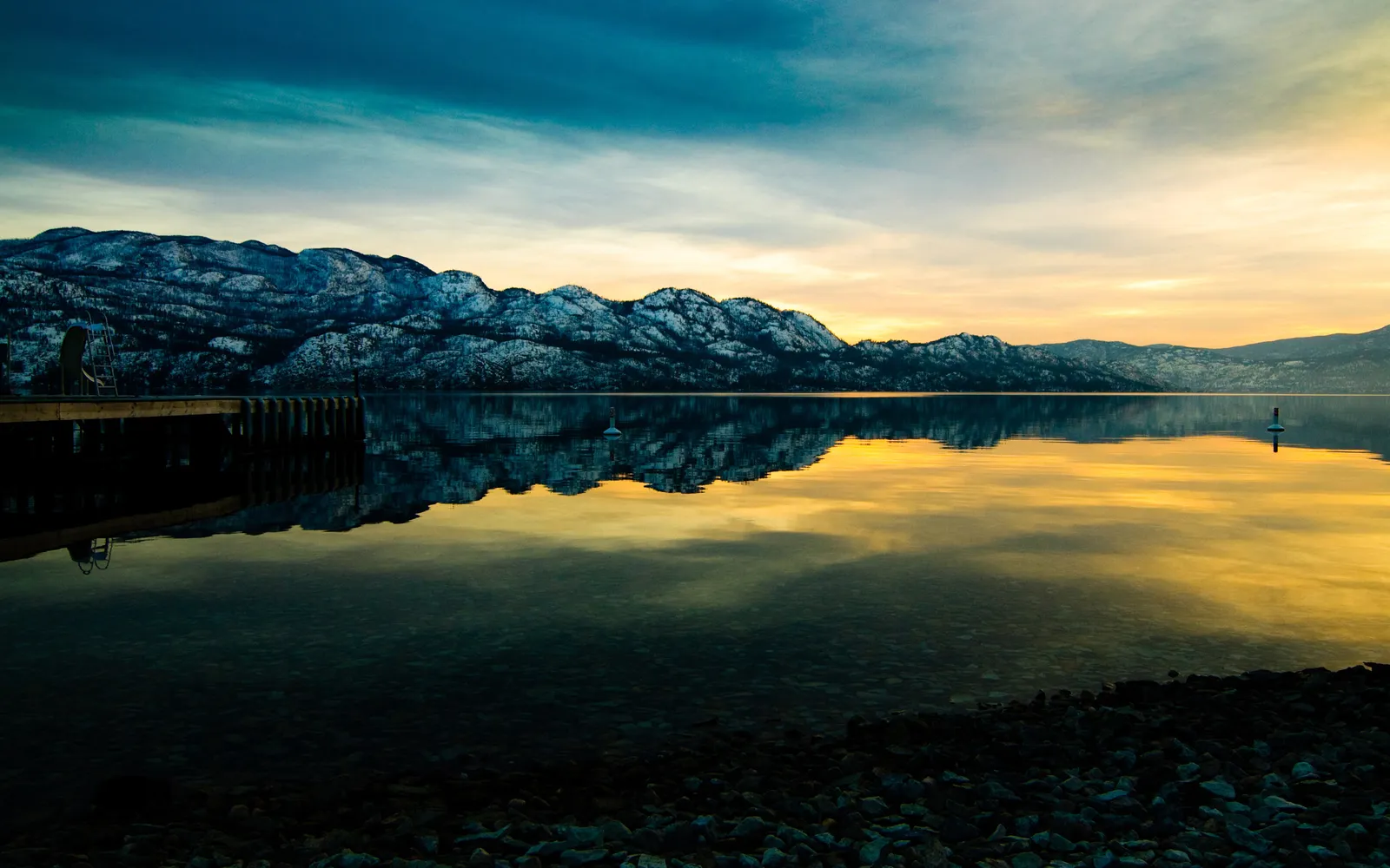 A calm lake reflects snow-capped mountains and a colorful sunset sky, with a wooden dock on the left.