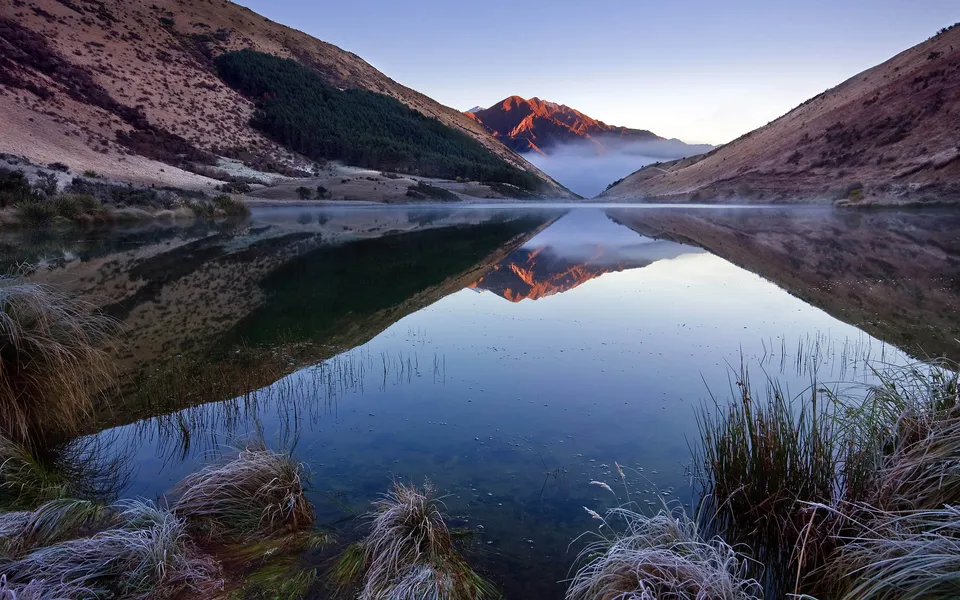 A lake reflects the sunrise colors on distant mountains, with mist hovering in the background.