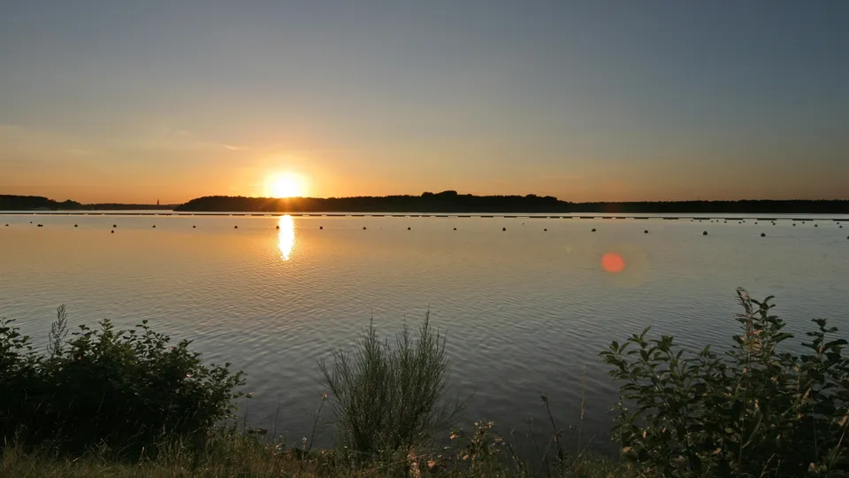 A serene sunset over a calm lake, with the sun low on the horizon and its reflection shimmering on the water.