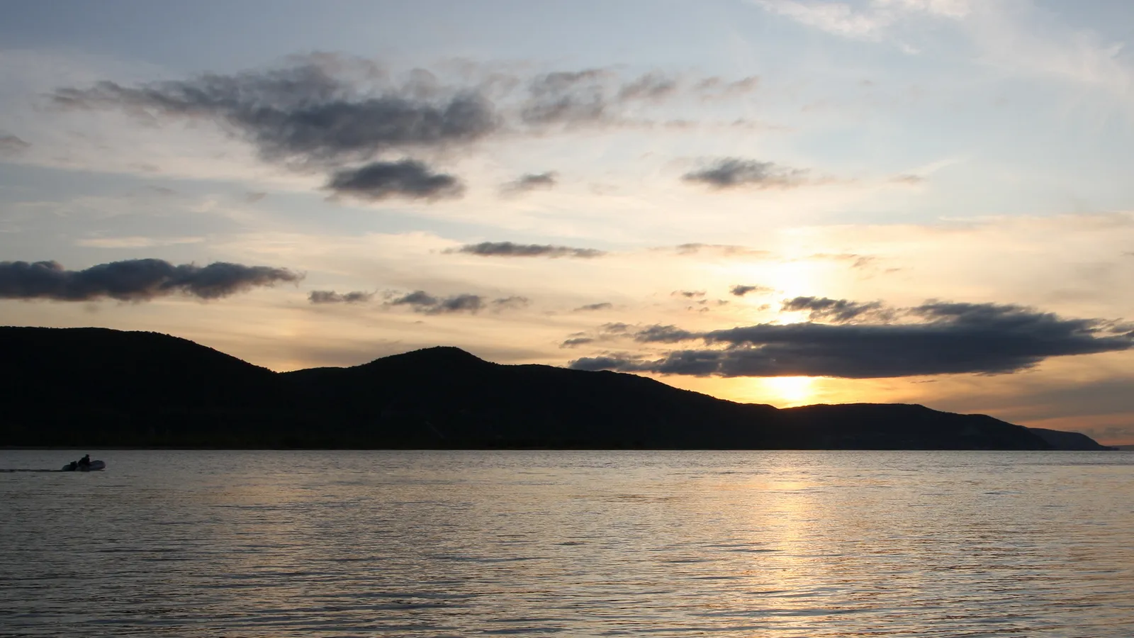A small boat travels across a calm lake towards silhouetted mountains under a colorful sunset sky.