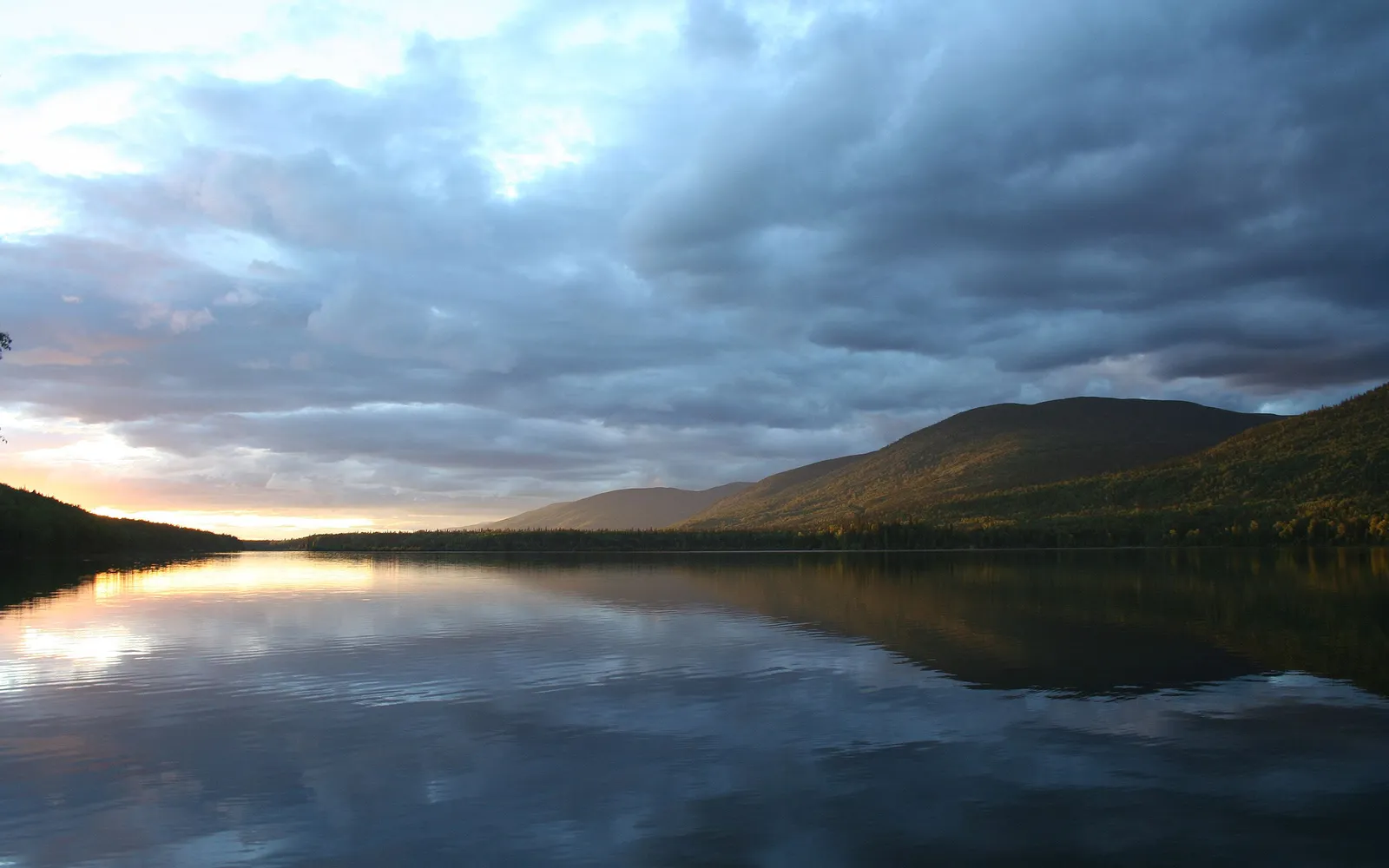 A wide lake reflects the sky and tree-covered mountains during a dramatic sunset.