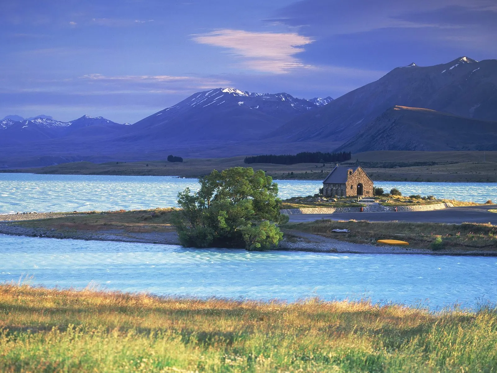 A small stone church sits on a small island in a bright blue lake, with mountains in the background.