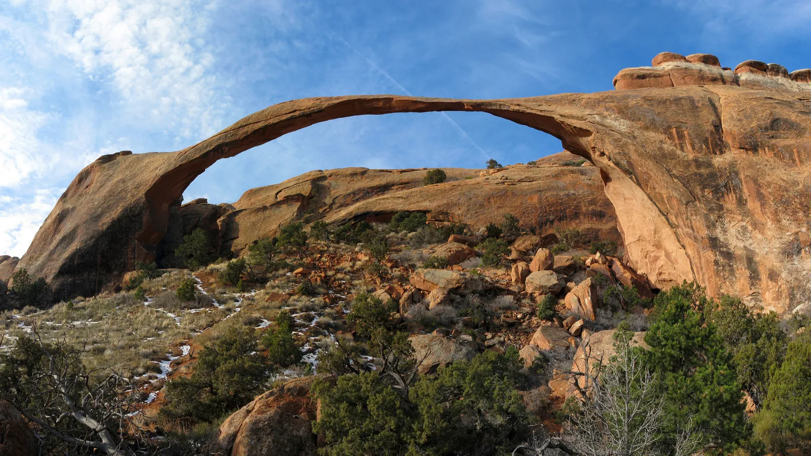 A wide, thin natural rock arch spans across a rocky desert landscape under a bright blue sky with wispy clouds.