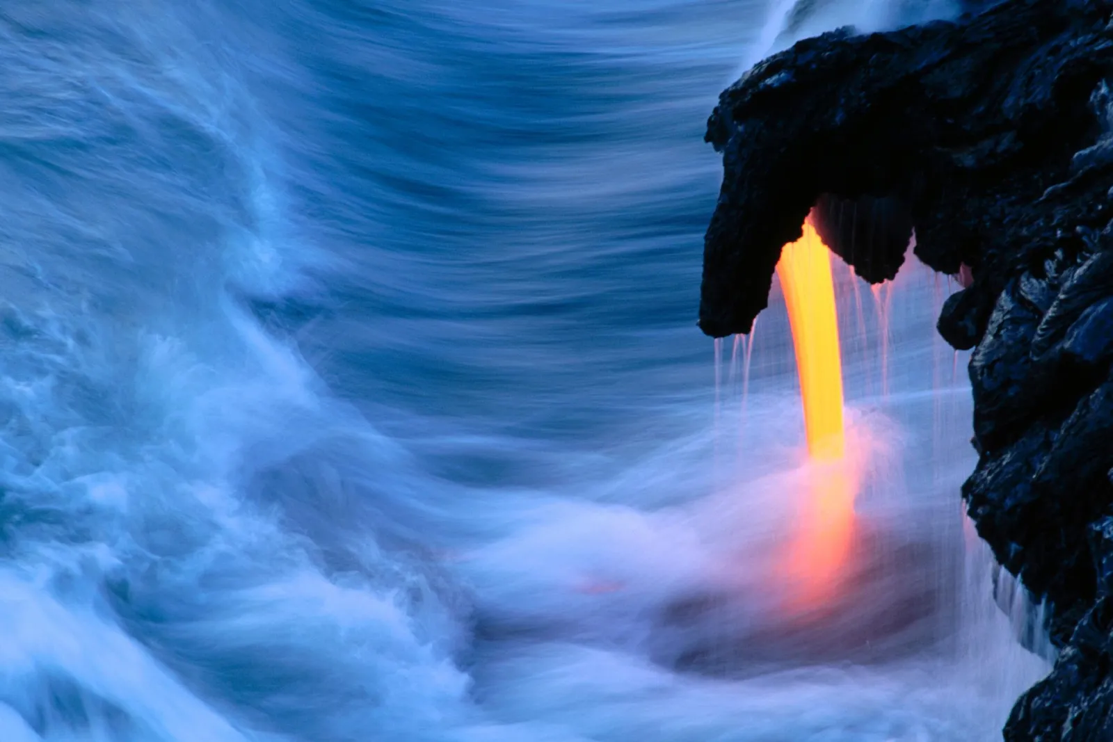 Molten orange lava streams down a dark rock cliff into the churning blue ocean.