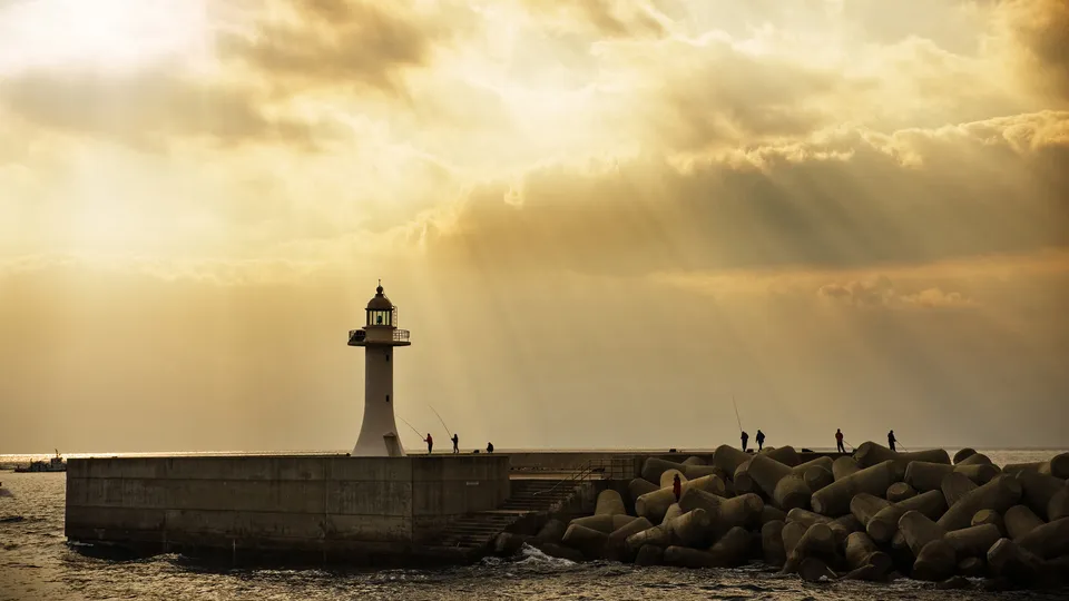 A lighthouse stands on a concrete breakwater with several people fishing, under a sky with dramatic sun rays.