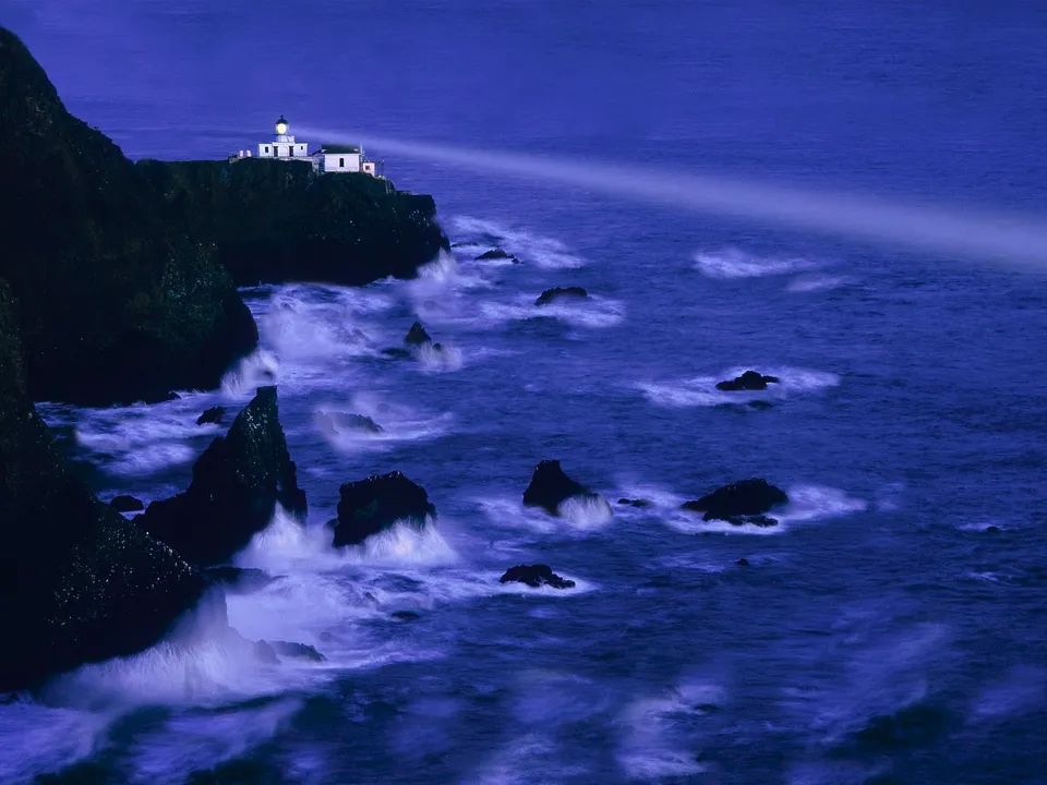 A lighthouse on a cliff shines its beam across the dark, choppy ocean at dusk.