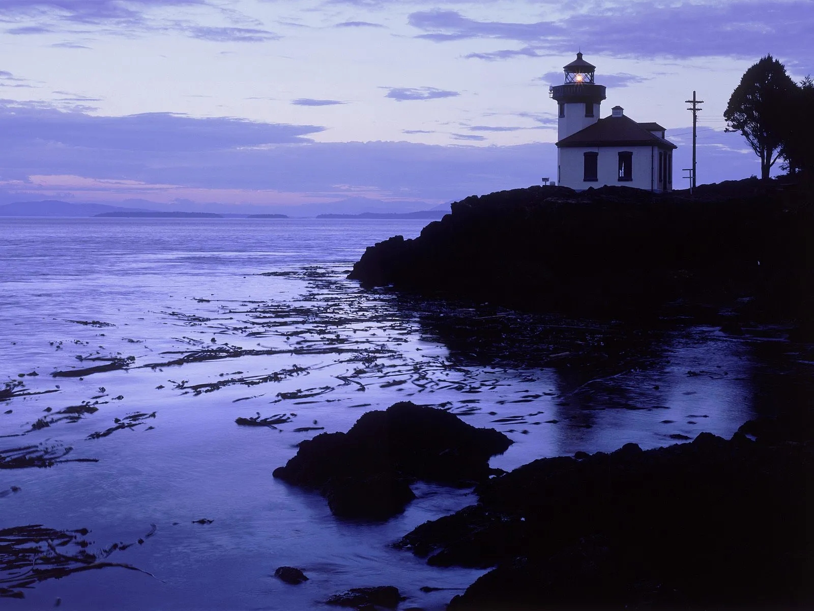 A lighthouse stands on a rocky coast at dusk, with the ocean stretching out to the horizon.