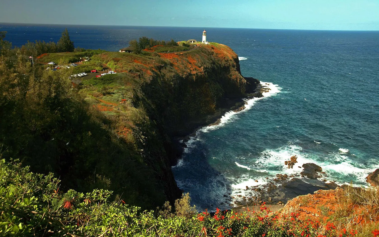 A white lighthouse stands on a grassy cliff overlooking the blue ocean with waves crashing on the rocks below.