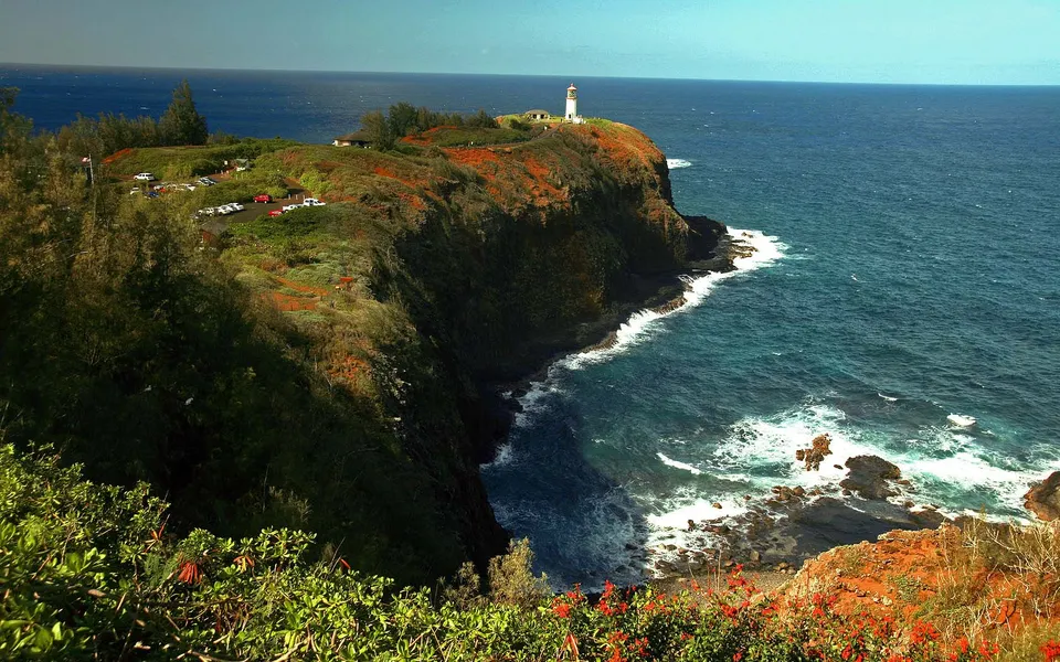 A white lighthouse stands on a grassy cliff overlooking the blue ocean with waves crashing on the rocks below.