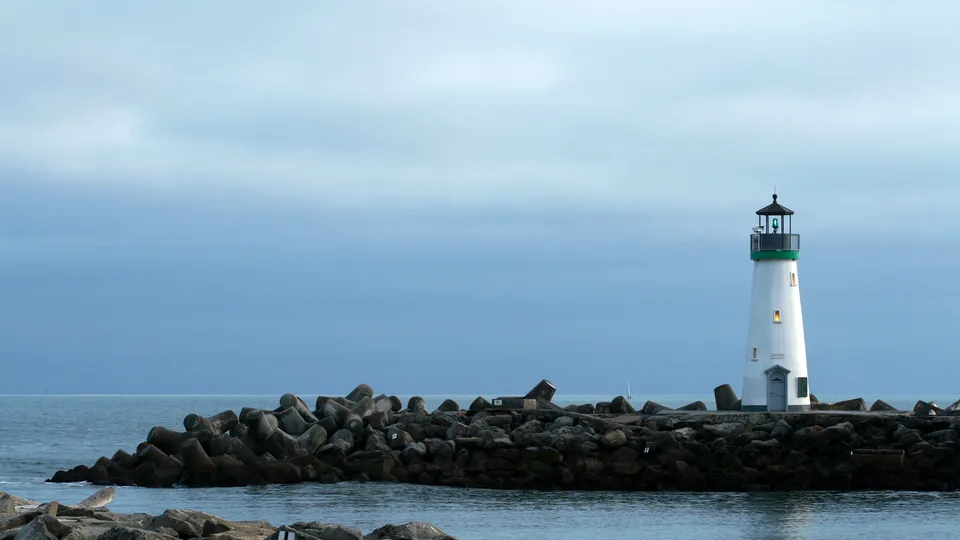 A white lighthouse with a green band stands on a rocky breakwater with the ocean and a cloudy sky in the background.
