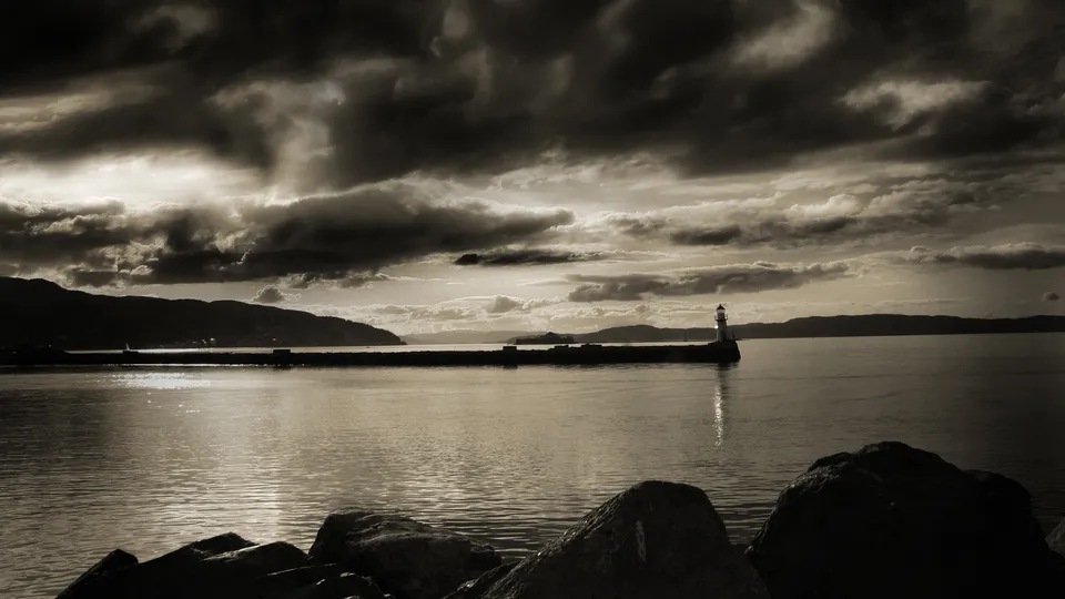 A lighthouse stands on a pier extending into the water under a dramatic, cloudy sky.