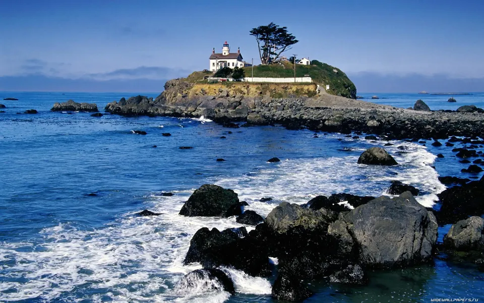 A white lighthouse stands on a grassy island surrounded by the blue ocean and rocky shores.