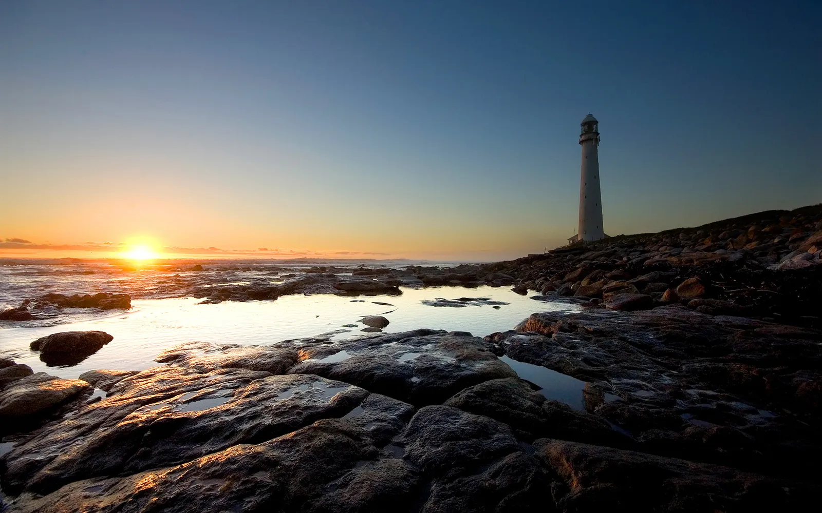 A tall white lighthouse stands on a rocky shore as the sun rises over the ocean.