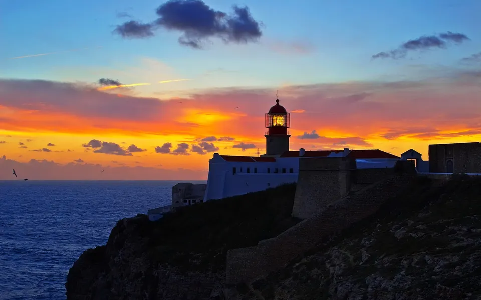 A lighthouse on a cliff overlooking the ocean during a vibrant sunset with orange and blue skies.