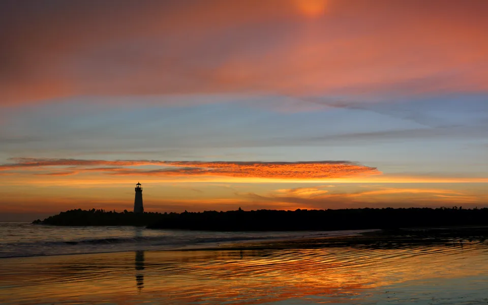 A lighthouse stands on a rocky outcrop against a vibrant sunset sky with colorful clouds reflected in the water.