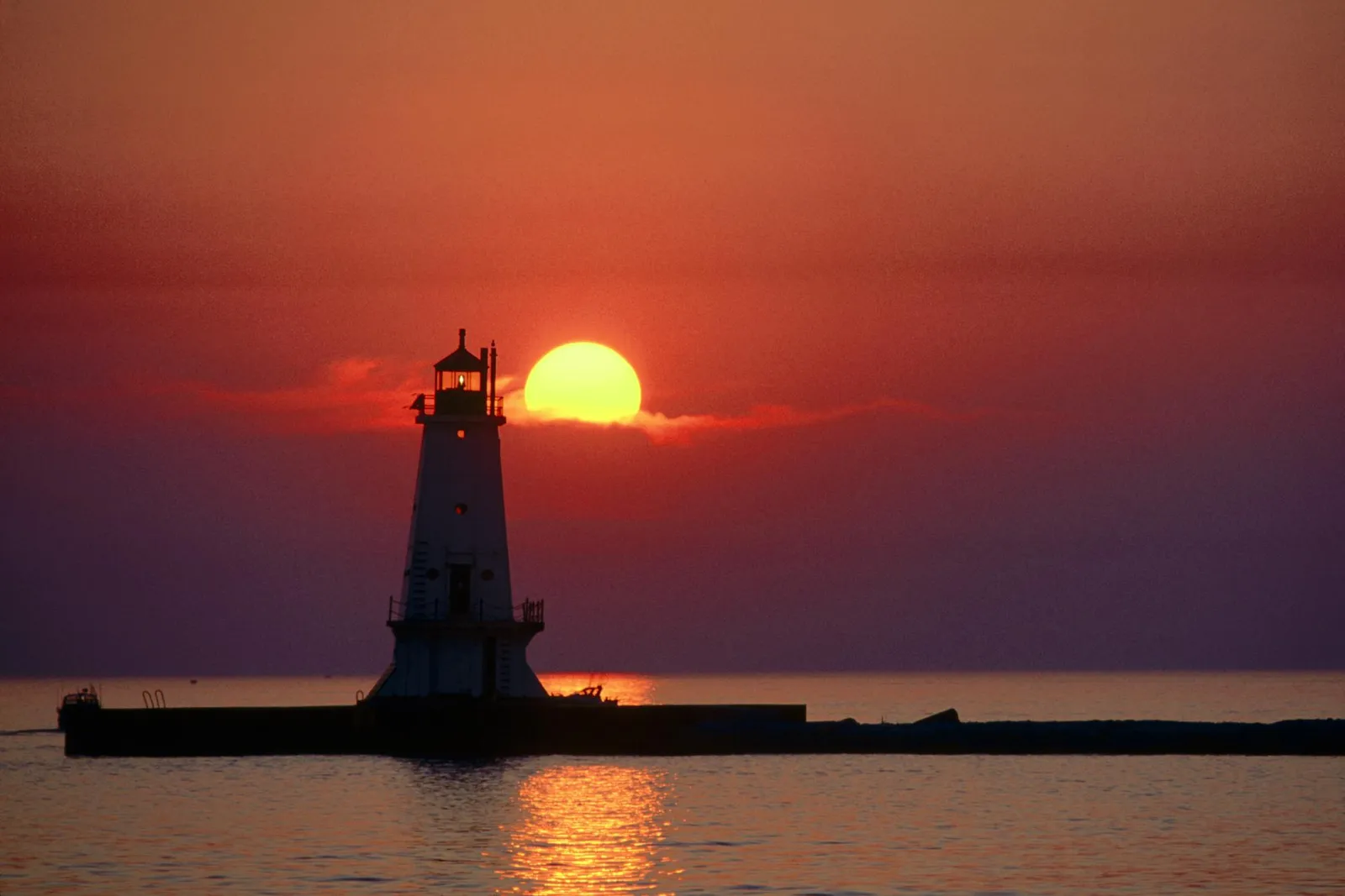 A white lighthouse stands on a pier as the sun sets behind it, casting a warm glow on the water.