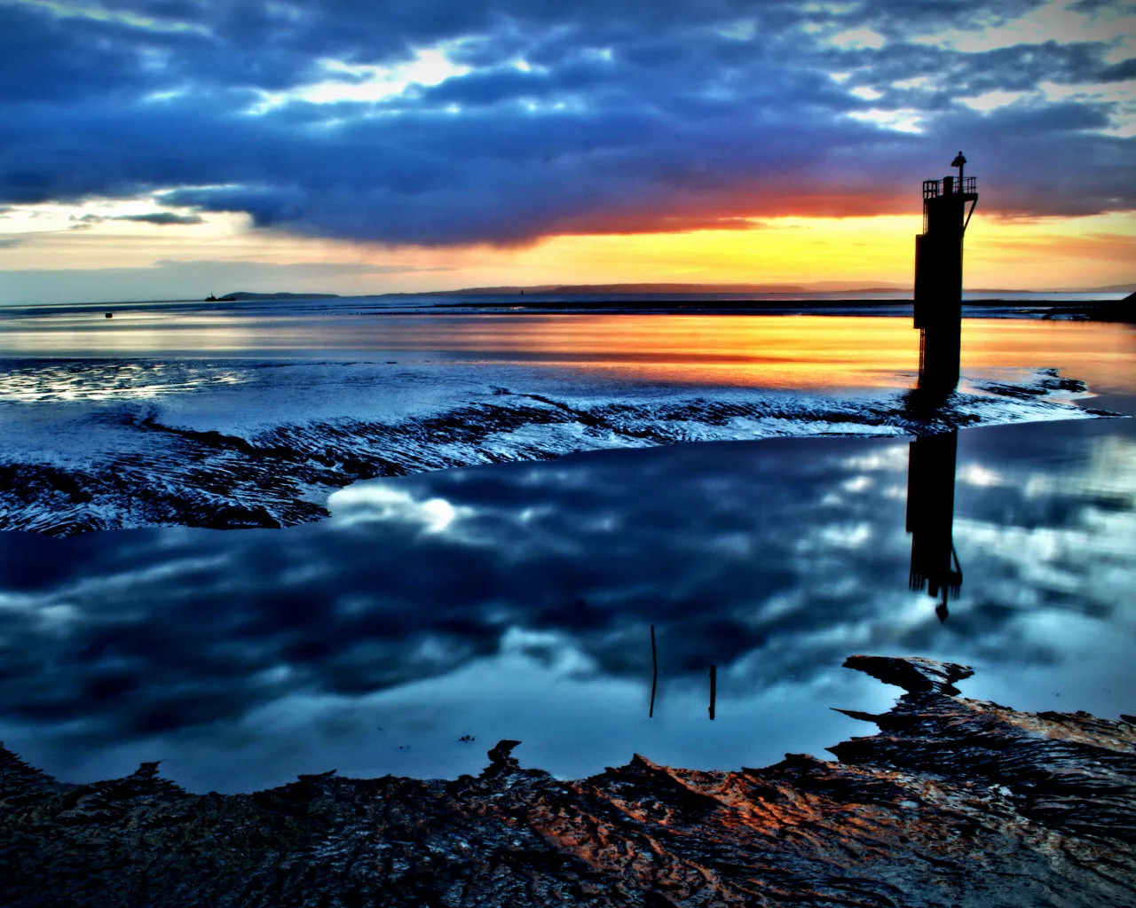 A silhouetted lighthouse stands on a tidal flat during a vibrant sunset, with clouds reflected in the water.