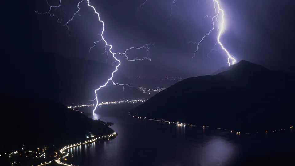 Lightning bolts illuminate the sky above a dark, mountainous landscape with a lake and city lights.