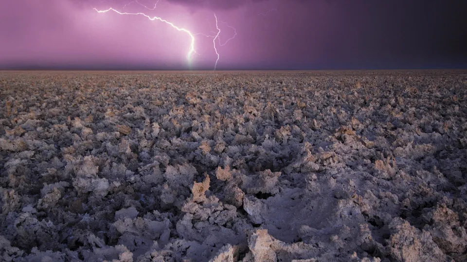 A bright lightning bolt strikes from a purple storm cloud over a desolate landscape of salt flats.