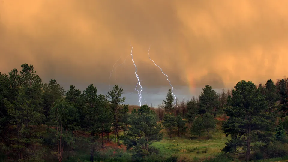 Lightning bolts strike down from a dramatic orange sky over a dark forest, with a faint rainbow visible.